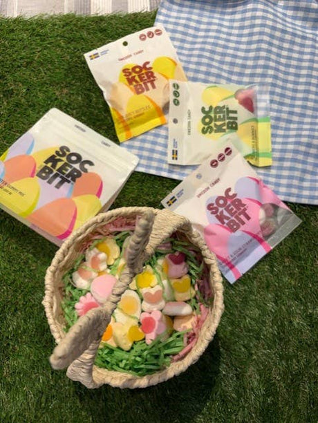 A woven basket of colorful gummies sits on green grass beside three open Sockerbit Sour Juicy Bottles Fruit Swedish Candy 2.8oz bags, with a blue and white checkered cloth nearby.