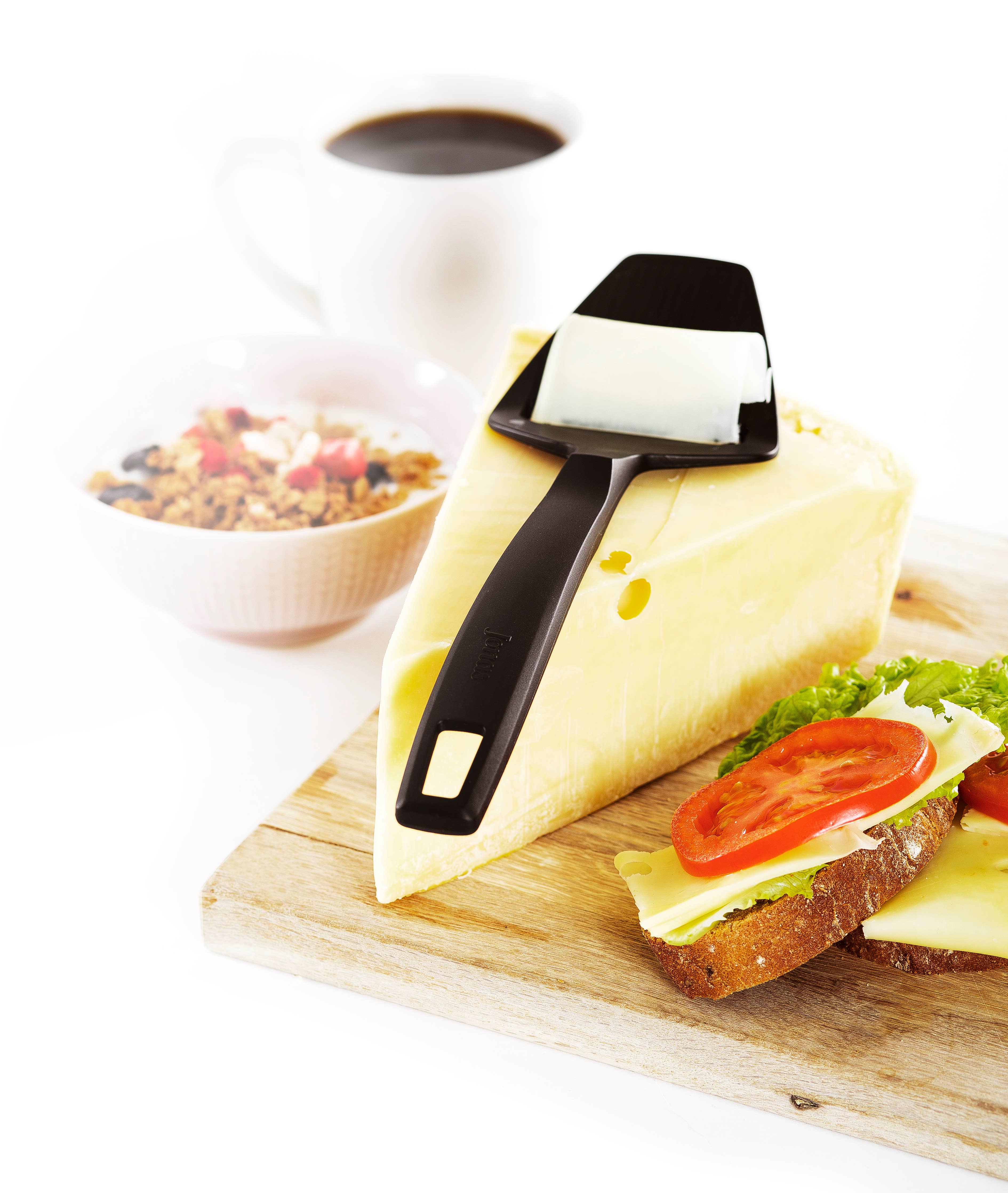 A Swedish Cheese Plane for Hard Cheese rests on a block of cheese beside an open sandwich layered with cheese, lettuce, and tomato. In the background, a steaming cup of coffee and a bowl of cereal are placed on a white surface.