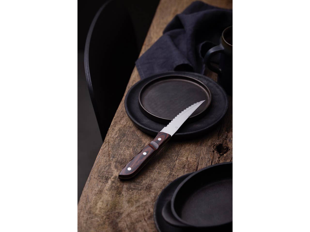 The Gense Old Farmer Steak Knife XL Classic, featuring a steel and wood design, rests on a dark plate alongside stainless steel cutlery on a rustic wooden table set with matching dark plates.