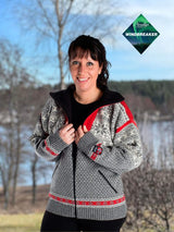 A woman stands outdoors, smiling at the camera in front of trees and water, wearing the Nordlys of Norway - Northern Lights windproof wool jacket. Windbreaker logo appears in the top right corner.