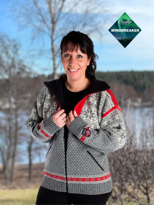 A person wearing the Nordlys of Norway Northern Lights Patterned windproof wool jacket for women and men stands outdoors against a snowy landscape backdrop, with the grey, natural, and red design adding a touch of magic to the scene.