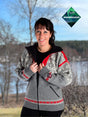 A woman stands outdoors, smiling at the camera in front of trees and water, wearing the Nordlys of Norway - Northern Lights windproof wool jacket. Windbreaker logo appears in the top right corner.