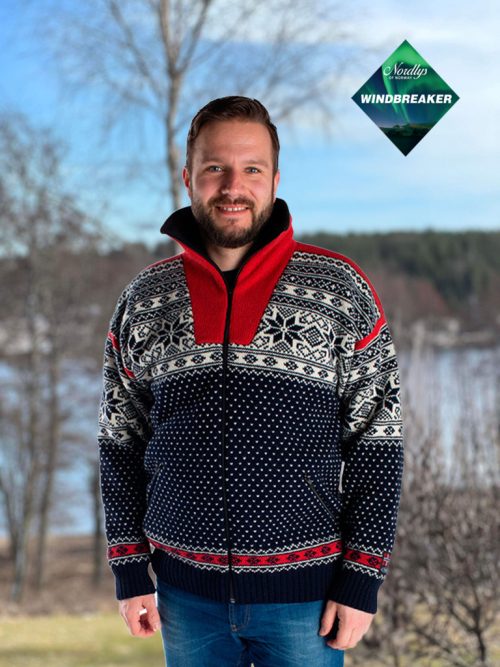 A person stands outdoors, smiling in a Nordlys of Norway sweater featuring a Northern Lights Patterned design. The sweater's red collar complements the natural backdrop of trees and water, while a green "Windbreaker" logo is displayed in the corner.