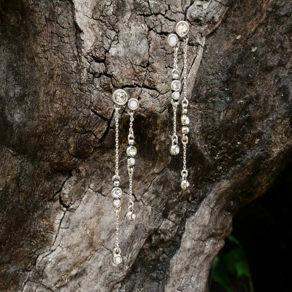 The A&C Oslo Long Cascade Crystal Chain Silver Earrings, featuring round white gemstones and delicate crystal chain accents, are displayed against the textured surface of a tree trunk.