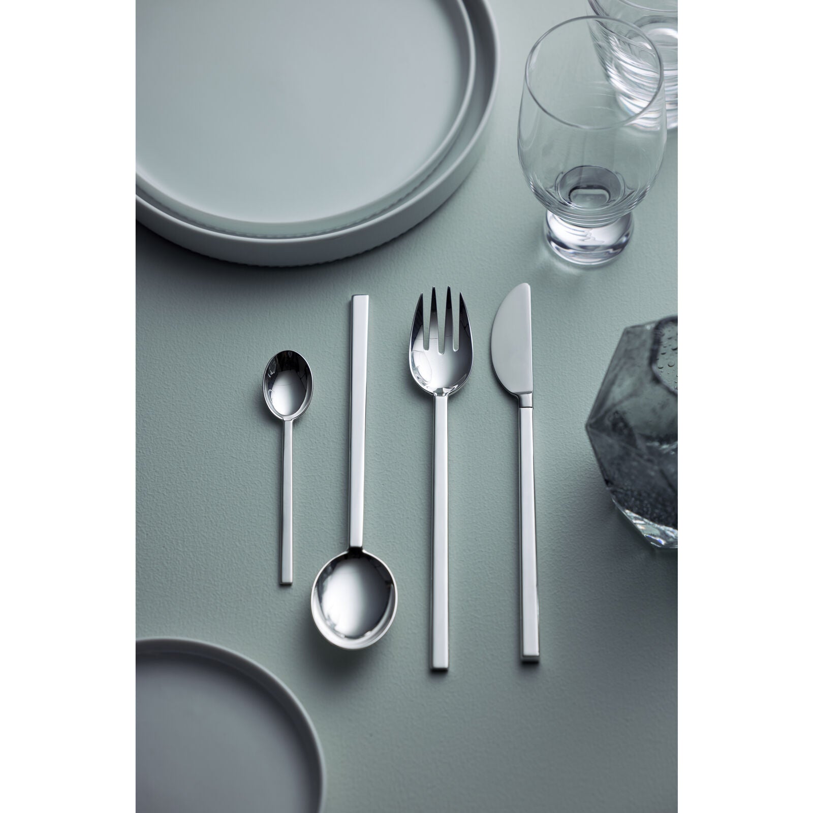 The Gense - Silver Dining Set CPB 2091 showcases premium cutlery, including a spoon, ladle, fork, and knife, all arranged neatly on a gray surface with adjacent plates and glassware.