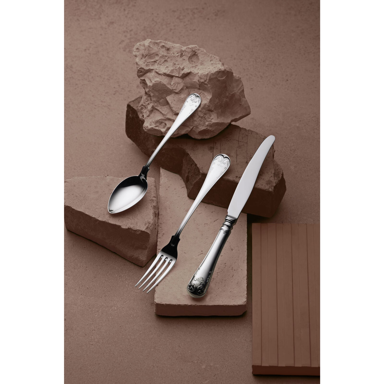 The Gense Silver Dining Set Gammal Fransk 830, featuring a spoon, fork, and knife with ornate handles, rests on textured stone blocks against a brown background.