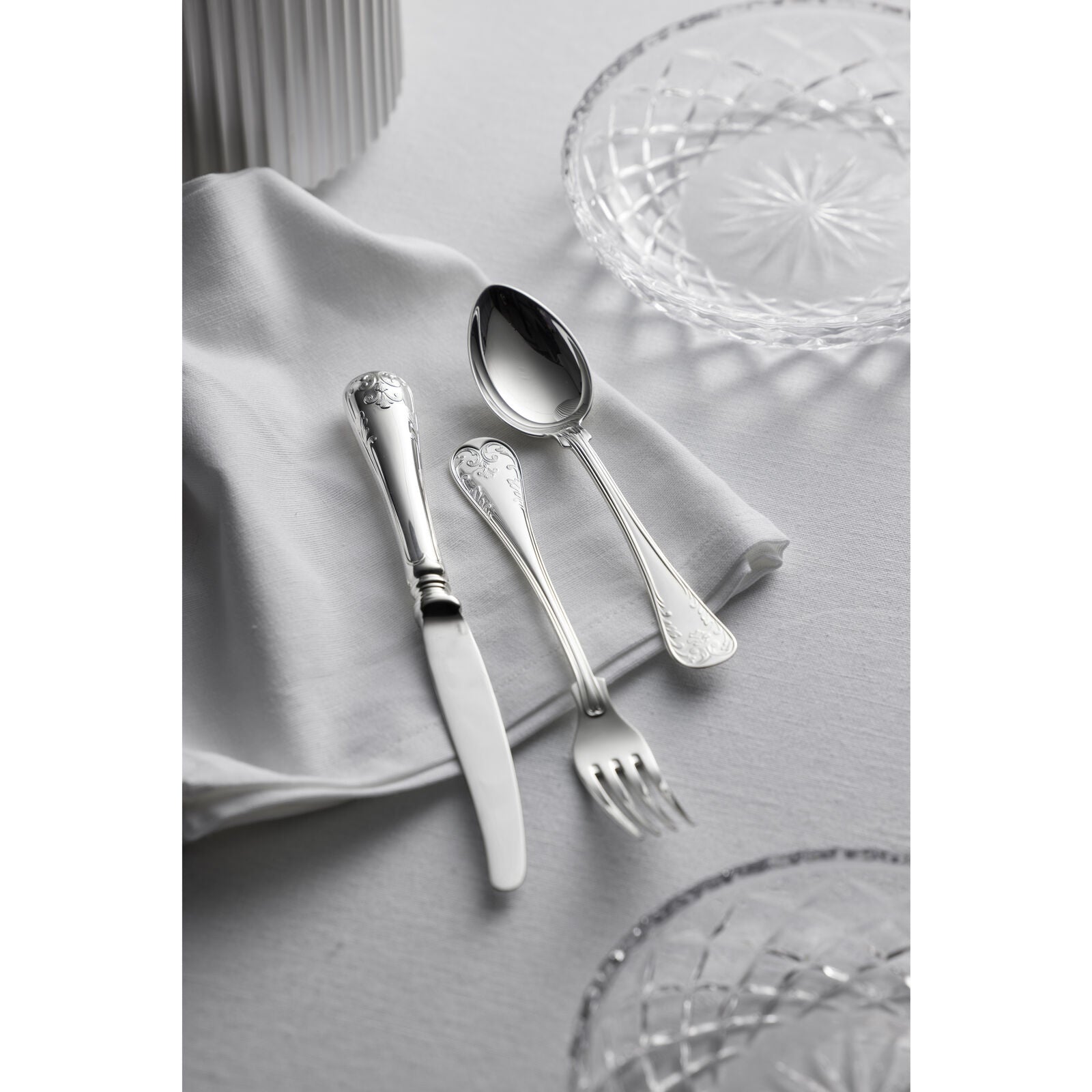 The Gense Silver Dining Set Gammal Fransk 830, featuring a knife, spoon, and fork, elegantly rests on a white cloth napkin, enhancing the look of two patterned glass plates on the pristine white tablecloth.