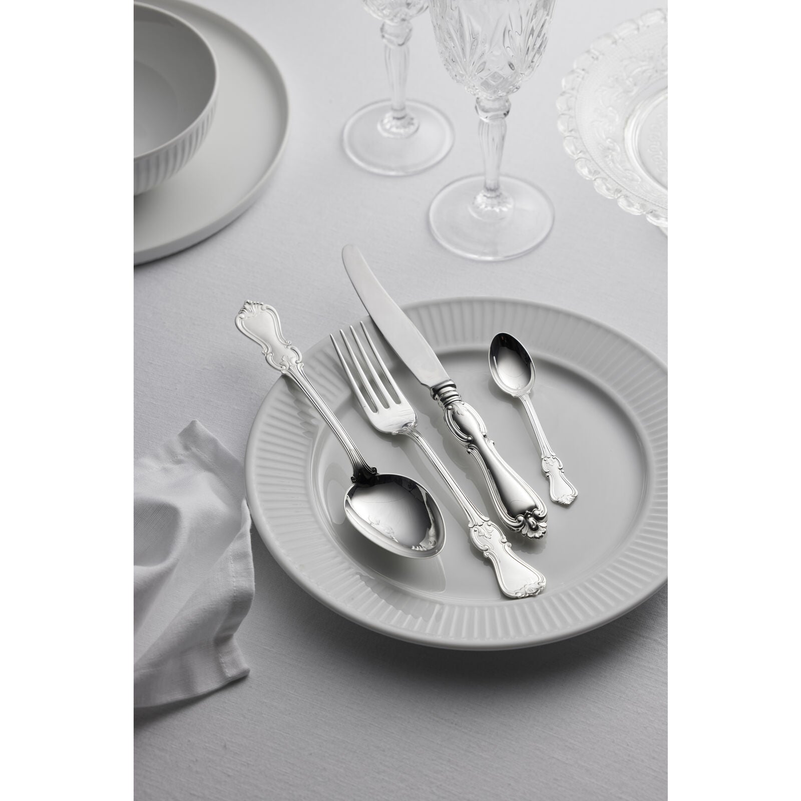 A white plate is adorned with the Gense Silver Dining Set Olga 830, featuring elegant Swedish silver knives, forks, and spoons. Nearby, crystal glasses sparkle beside a neatly folded napkin on a pristine white tablecloth.