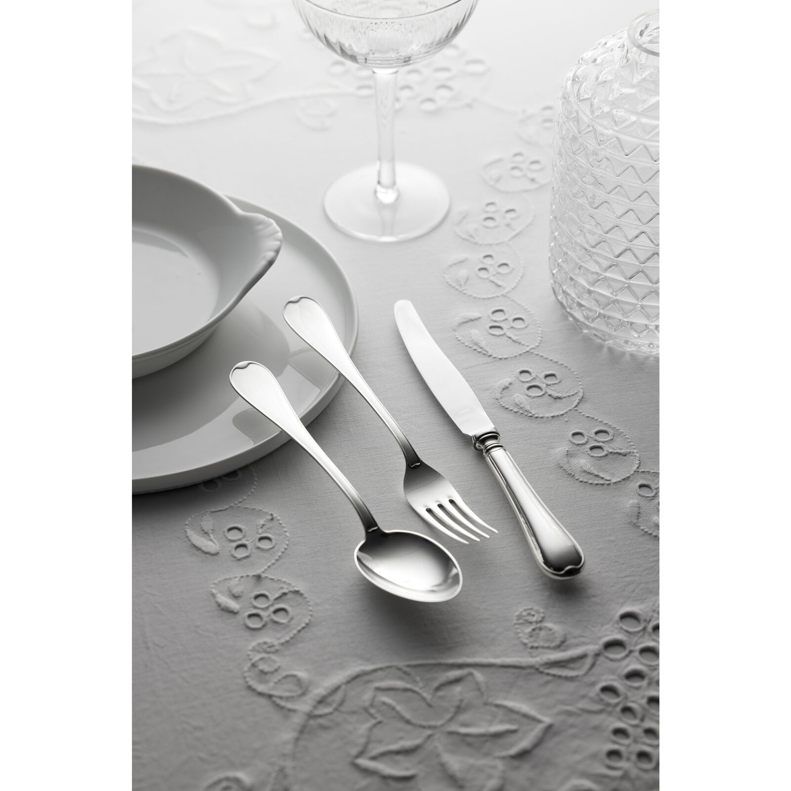 The Gense - Silver Dining Set Svensk 830, made in Sweden and featuring a fork, knife, and spoon, is elegantly arranged on a floral white tablecloth, partnered with a white dish and wine glass.