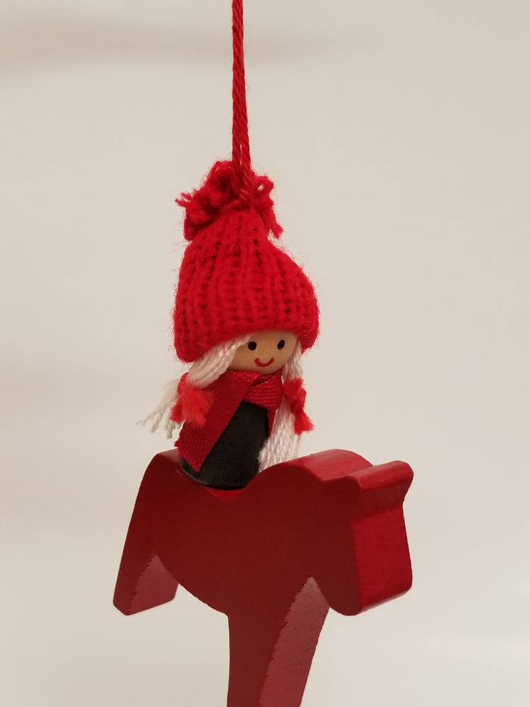 A charming Christmas tree ornament features a girl wearing a red knit hat, riding a red Swedish Dala horse, all suspended by a red string against a plain background.