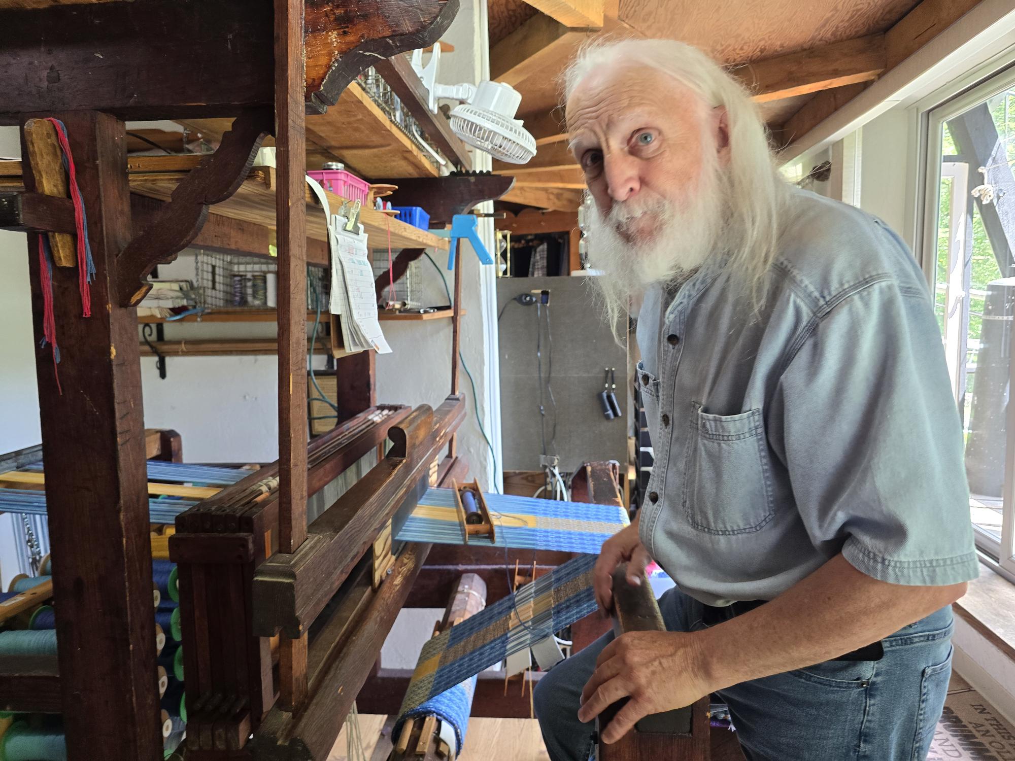 An elderly person with long white hair meticulously weaves blue and white threads on a wooden loom indoors, crafting a Runner: Swedish Flag Runner (Length 46"-49") from 100% cotton.