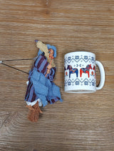 A ceramic mug with a Nordic horse pattern sits next to the Philippa Blue Kitchen Witch Doll, handmade in Germany, on a wooden surface.
