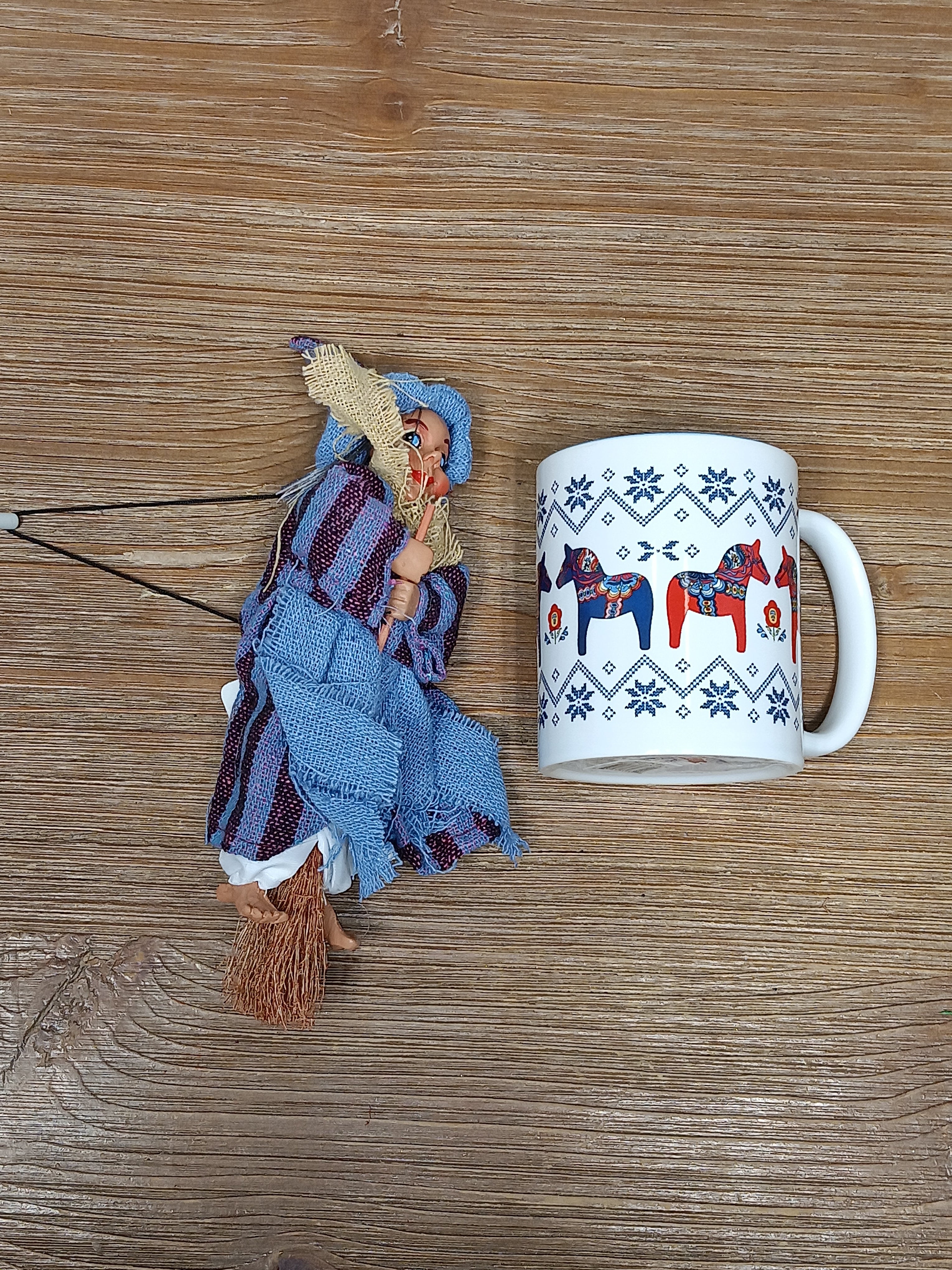 A ceramic mug with a Nordic horse pattern sits next to the Philippa Blue Kitchen Witch Doll, handmade in Germany, on a wooden surface.