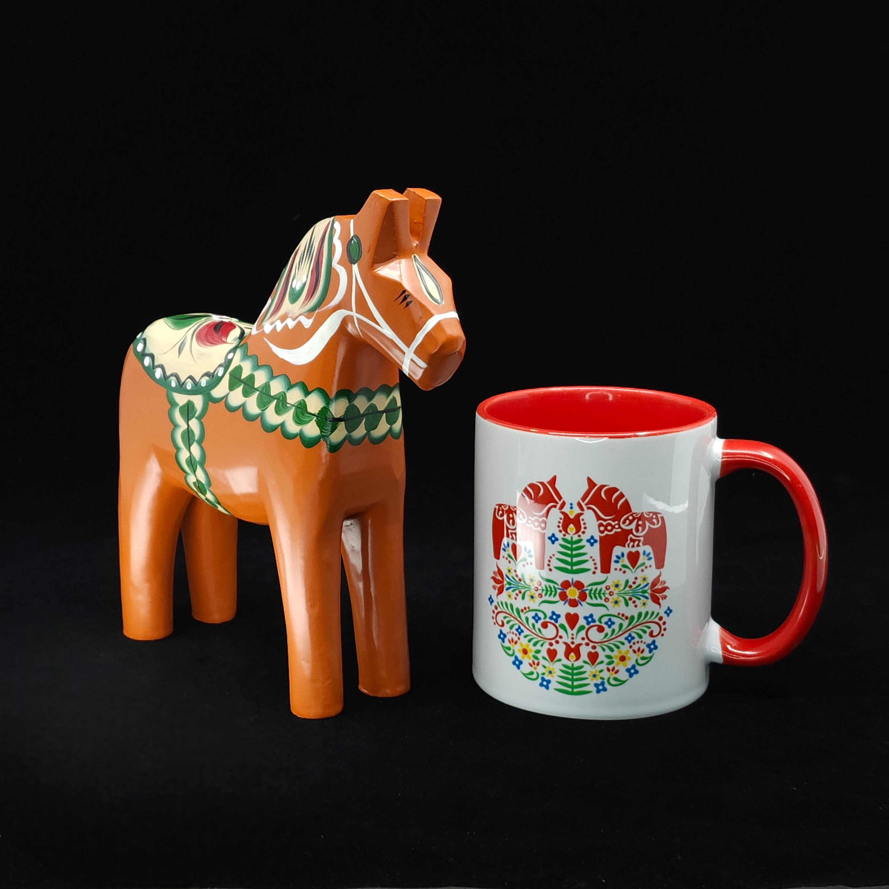 An antique-inspired Swedish Dala Horse in red-orange (17cm) stands beside a white mug with a red handle and colorful folk art pattern, both displayed against a black background.