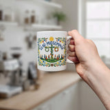 A hand holds the Swedish Gift Store’s "Glad Midsommar" 11 oz mug, featuring colorful florals and maypole dancers with GLAD MIDSOMMAR text; a blurred kitchen appears in the background.