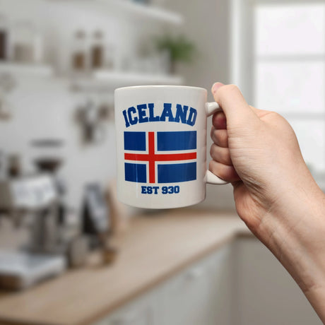 A hand holds the Swedish Gift Store Mug: Established Series Iceland Flag Mug with Year Founded (11 oz), featuring “Iceland,” the national flag, and “EST 930,” in a bright, modern kitchen.