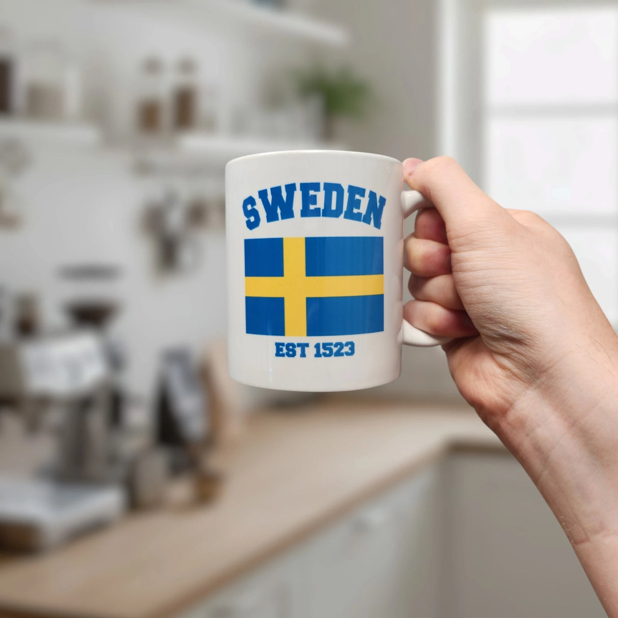 A hand holds the Swedish Gift Store's 11oz Established Series Sweden Flag Mug featuring the blue and yellow flag, "SWEDEN," and "EST 1523." A bright, modern kitchen is blurred in the background.