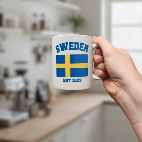 A hand holds the Swedish Gift Store's 11oz Established Series Sweden Flag Mug featuring the blue and yellow flag, "SWEDEN," and "EST 1523." A bright, modern kitchen is blurred in the background.