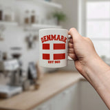 A hand holds the Swedish Gift Store's Mug: Established Series Denmark Flag Mug with Year Founded (11 oz), featuring "DENMARK," a red cross flag, and "EST 965" on it. A blurred kitchen with shelves, plants, and a window is in the background.