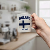 A person holds the Swedish Gift Store's Mug: Established Series Finland Flag Mug with Year Founded (11 oz), displaying “FINLAND EST 1917,” against a blurred kitchen—ideal as a Finnish coffee mug or heritage gift.