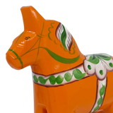 Close-up of an orange Swedish Dala horse made of pine wood, decorated with green and white painted patterns, shown on a white background.