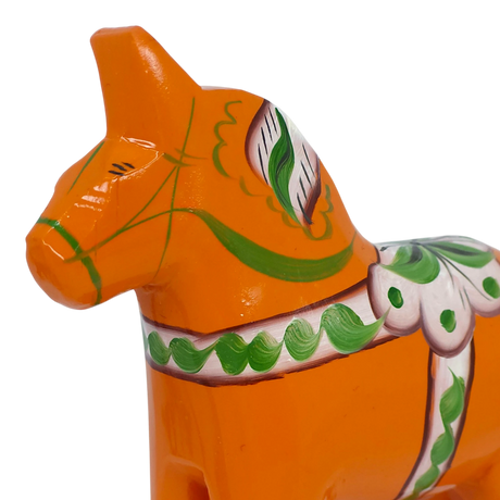 Close-up of an orange Swedish Dala horse made of pine wood, decorated with green and white painted patterns, shown on a white background.