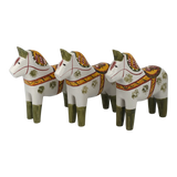 Three Dala horses with green legs, yellow and red saddles, and floral designs are arranged in a row facing left on a plain background.