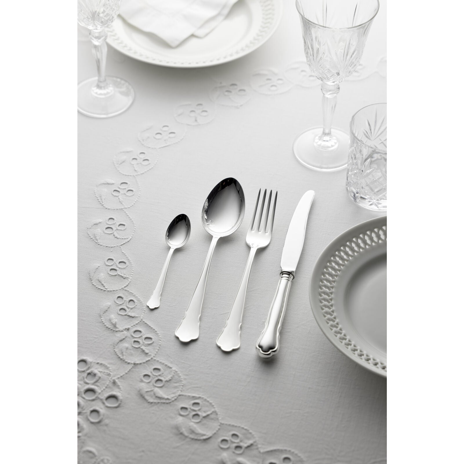 The sophisticated Gense - Silver Dining Set Chippendale 830, including a spoon, fork, knife, and small spoon, is beautifully displayed on a white embroidered tablecloth with immaculate white plates and shining crystal glasses.