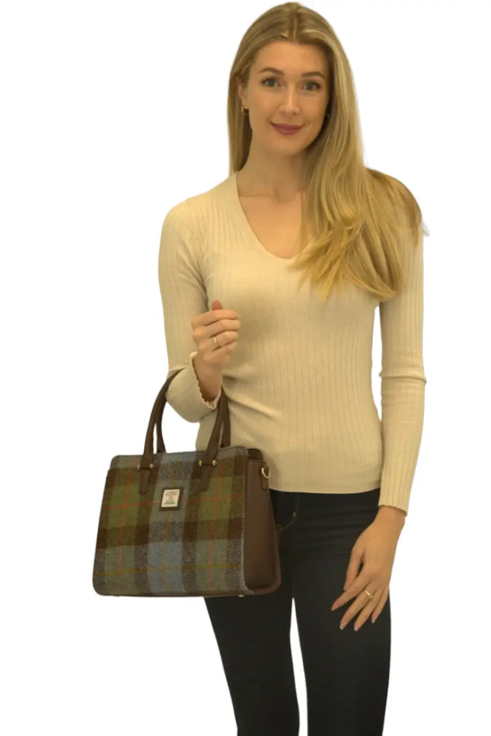 A woman stands against a white background in a beige sweater and dark pants, holding the Harris Tweed Double Handle Tote - Dark Brown with double handles and brown accents in her left hand.