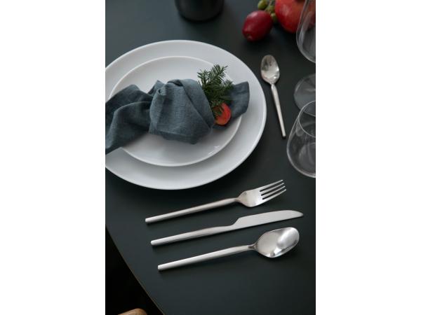 A neatly set dining table showcases a white plate, blue napkin, and herbs, enhanced by the modern classic Gense: Dorotea Cutlery Set in Matte Steel on the right. Shimmering empty glasses stand alongside a vibrant red pepper.