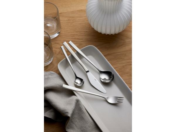 The Gense: Fuga Cutlery Set - Matte/Polished Steel, consisting of a knife, spoon, and fork, is elegantly arranged on a rectangular white plate on a wooden table. Nearby are two empty glasses with a white vase and a folded gray napkin.