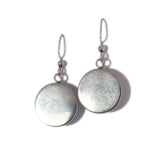 The "Mushrooms" earrings by Sarah Angst feature handmade silver round discs with hooks, gleaming against a white background and showcasing the artisan's craftsmanship.