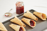 Six waffle cones filled with red jam, shaped perfectly with the Krumkake Cone Roller (Beechwood, 1.5" x 1.5" x 8.25"), rest on a tray beside an open jar of jam and a spoon on a white surface.