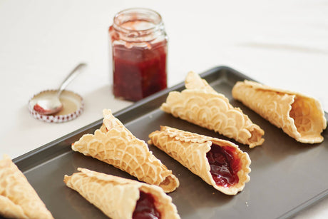 Six waffle cones filled with red jam, shaped perfectly with the Krumkake Cone Roller (Beechwood, 1.5" x 1.5" x 8.25"), rest on a tray beside an open jar of jam and a spoon on a white surface.