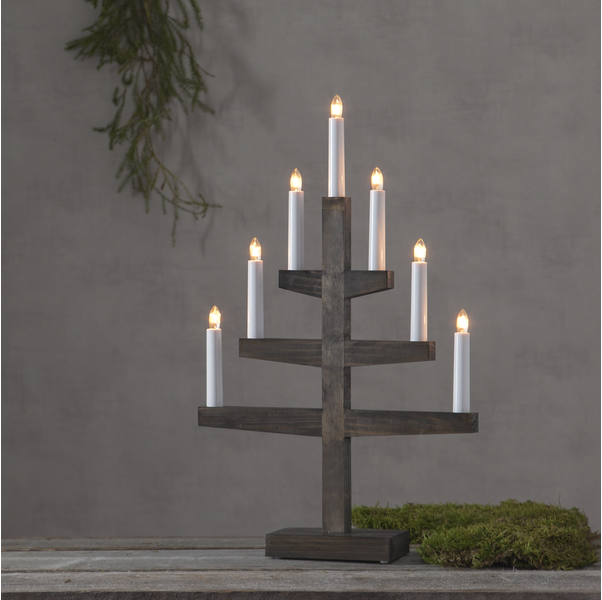 The Star Trading Trapp 7 Light Candelabra in walnut stain sits on a moss-adorned wooden table, evoking Christmas warmth against a gray wall.