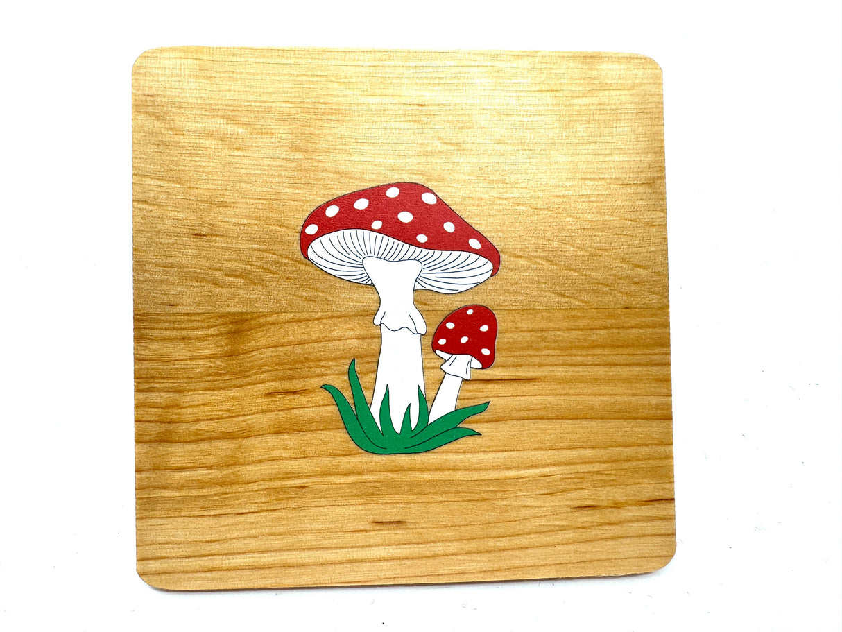 A handmade alder wooden coaster featuring two red mushrooms with white spots is set against a rustic wooden board. Made in Estonia, the product is named Coaster: Mushrooms on Alder Wood Coaster.