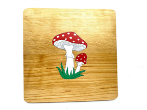 A handmade alder wooden coaster featuring two red mushrooms with white spots is set against a rustic wooden board. Made in Estonia, the product is named Coaster: Mushrooms on Alder Wood Coaster.