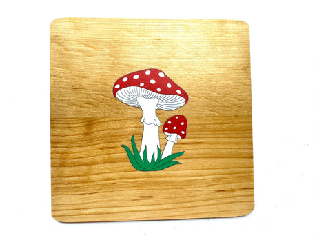 A handmade alder wooden coaster featuring two red mushrooms with white spots is set against a rustic wooden board. Made in Estonia, the product is named Coaster: Mushrooms on Alder Wood Coaster.