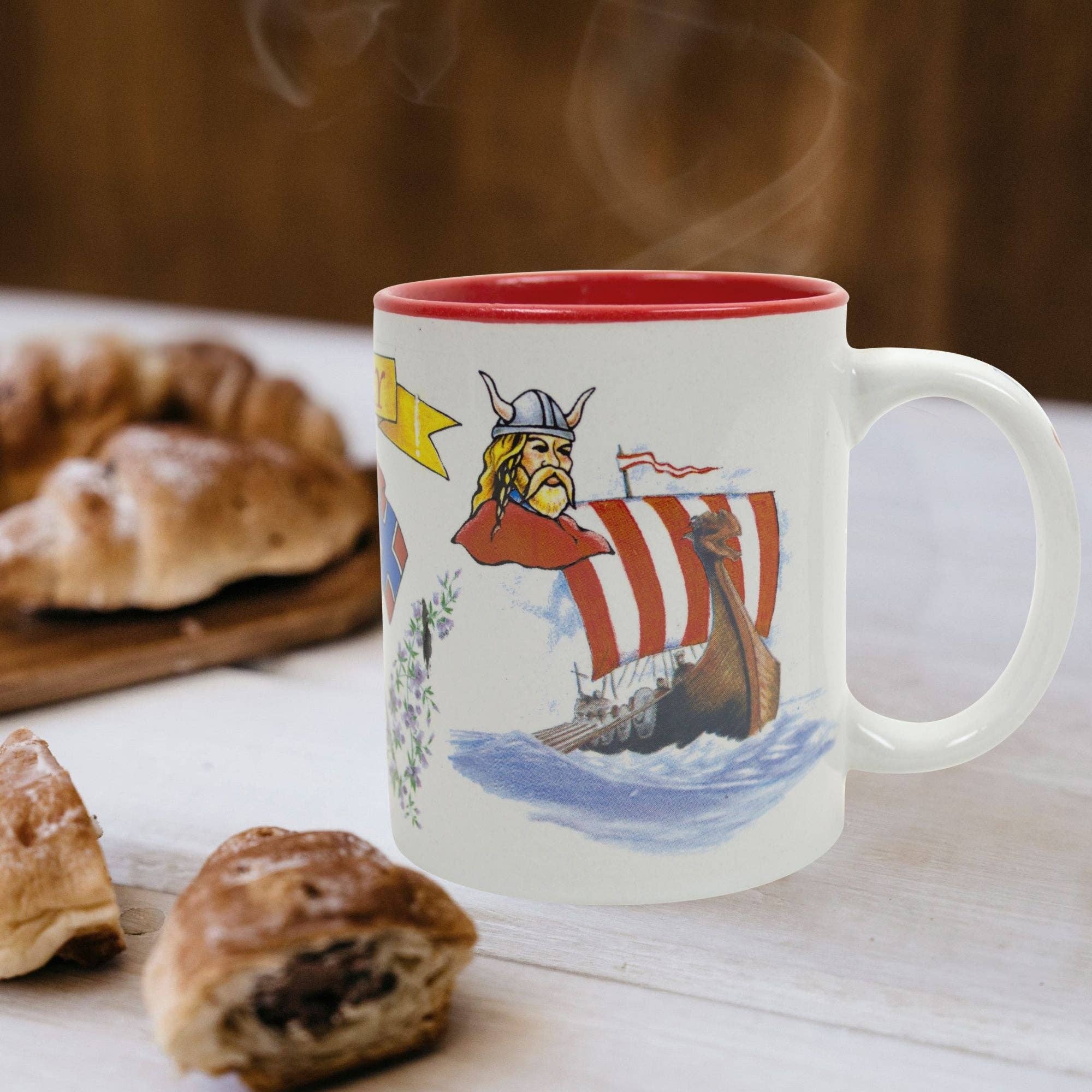 The I Love Norway coffee mug, featuring a Viking face and ship with intricate Norwegian graphics, sits on a table surrounded by delicious pastries.