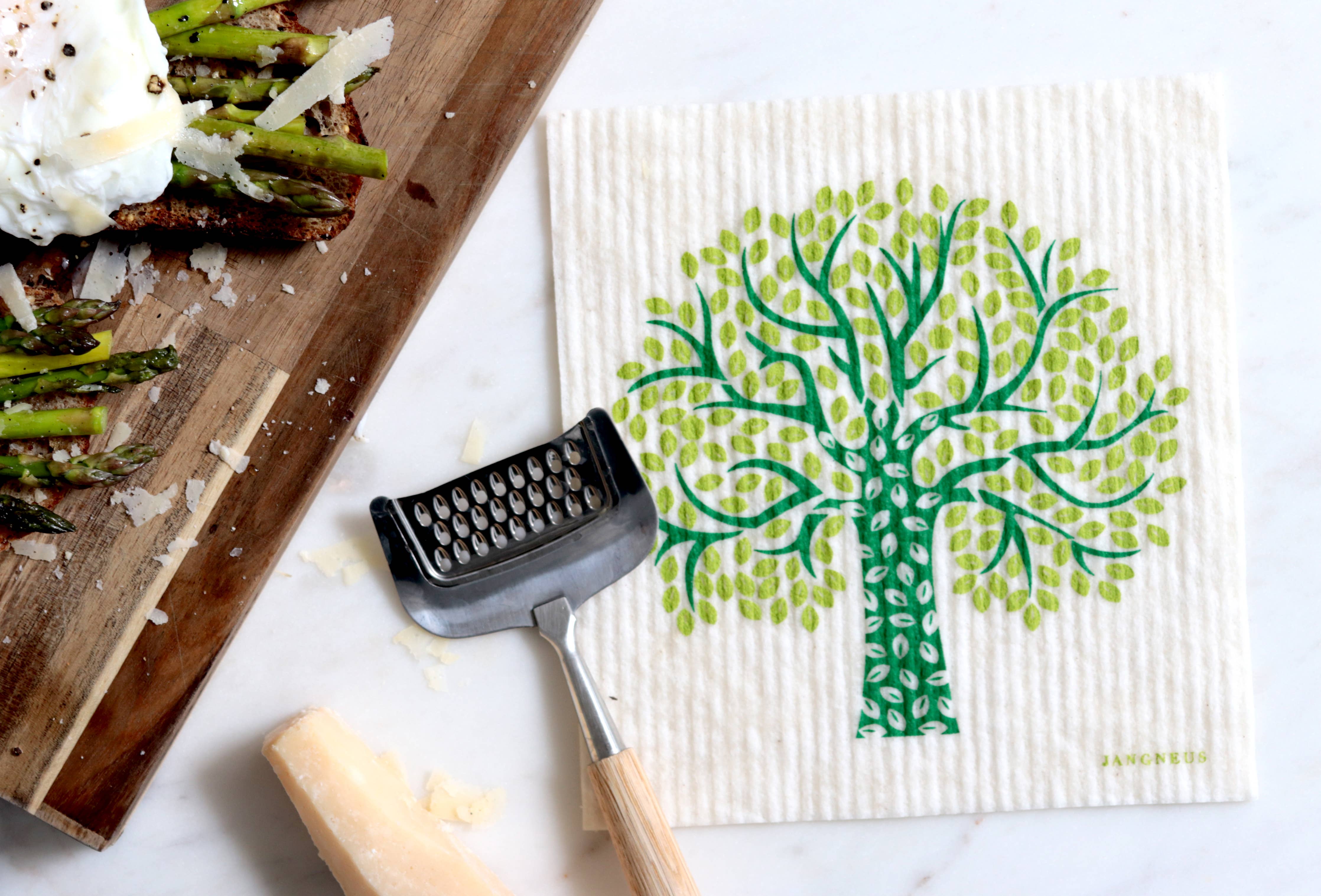 A Scandinavian style kitchen features a wooden board with asparagus-topped toast, a cheese grater, and a block of cheese. Nearby, Dish Cloths in the Tree - Green design complement the scene with both charm and practicality.