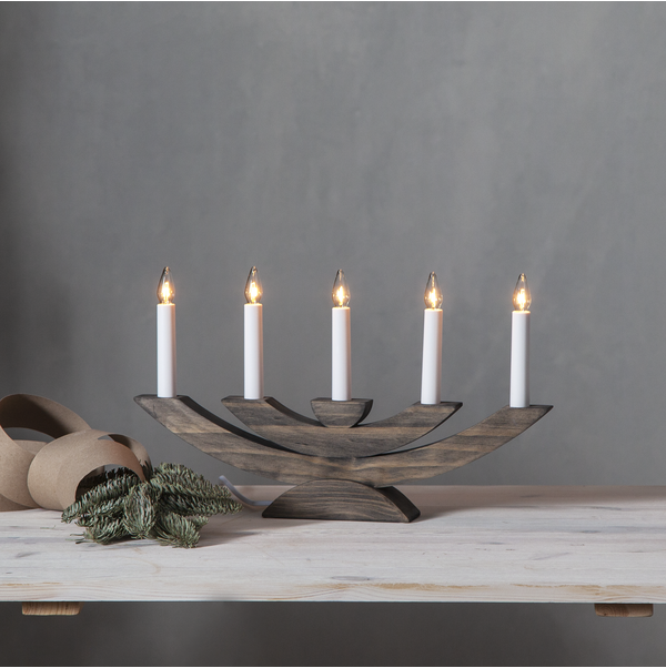 The "Candelabra: Star Trading Navida Candlestick Brown" with five lit candles sits on the table, casting a warm glow. Pine branches and a roll of brown paper lie beside it, creating a cozy Christmas atmosphere.