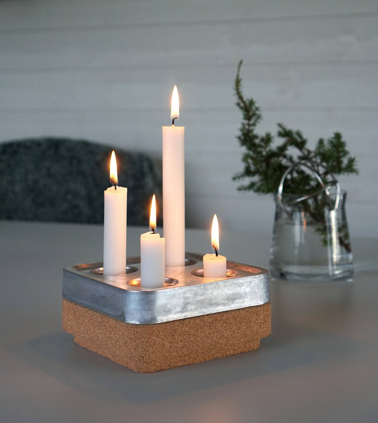 The Stumpastaken Fyran Four Candle Holder—square, made from 100% recycled white powder-coated material—features four white candles of varying heights, adding sustainable Swedish style to your decor. A glass vase with greenery sits in the background.
