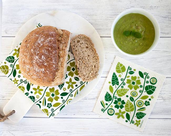 A round loaf of bread, with a slice cut, sits on a white plate next to a bowl of comforting green soup atop floral-patterned cloth and napkin on a white wooden surface, evoking thoughts of Melted Cheese: 60 Gorgeously Gooey Recipes.