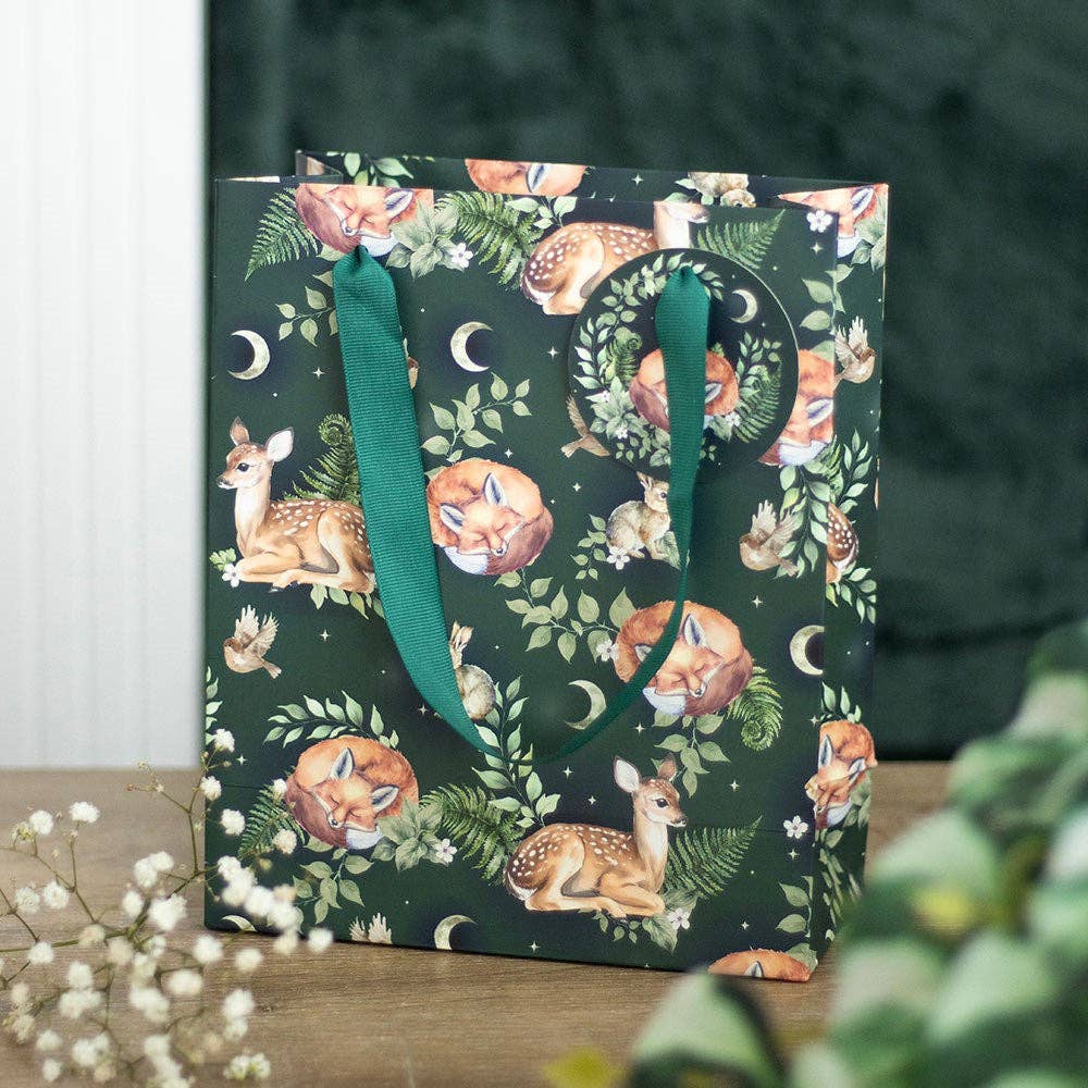 The Gift Bag: Fawn and Friends Forest Animal Print Medium Gift Bag with green ribbon handles, featuring deer, foxes, ferns, and crescent moons, rests on a wooden surface.