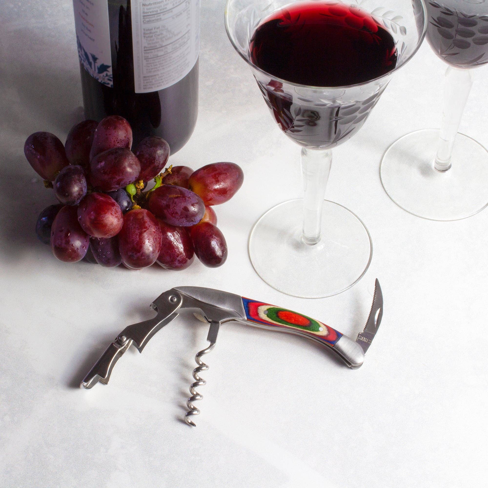 A wine glass and bottle sit elegantly beside a bunch of grapes, accompanied by a corkscrew and the Marrakesh Collection Waiter's Friend Corkscrew on a light surface.