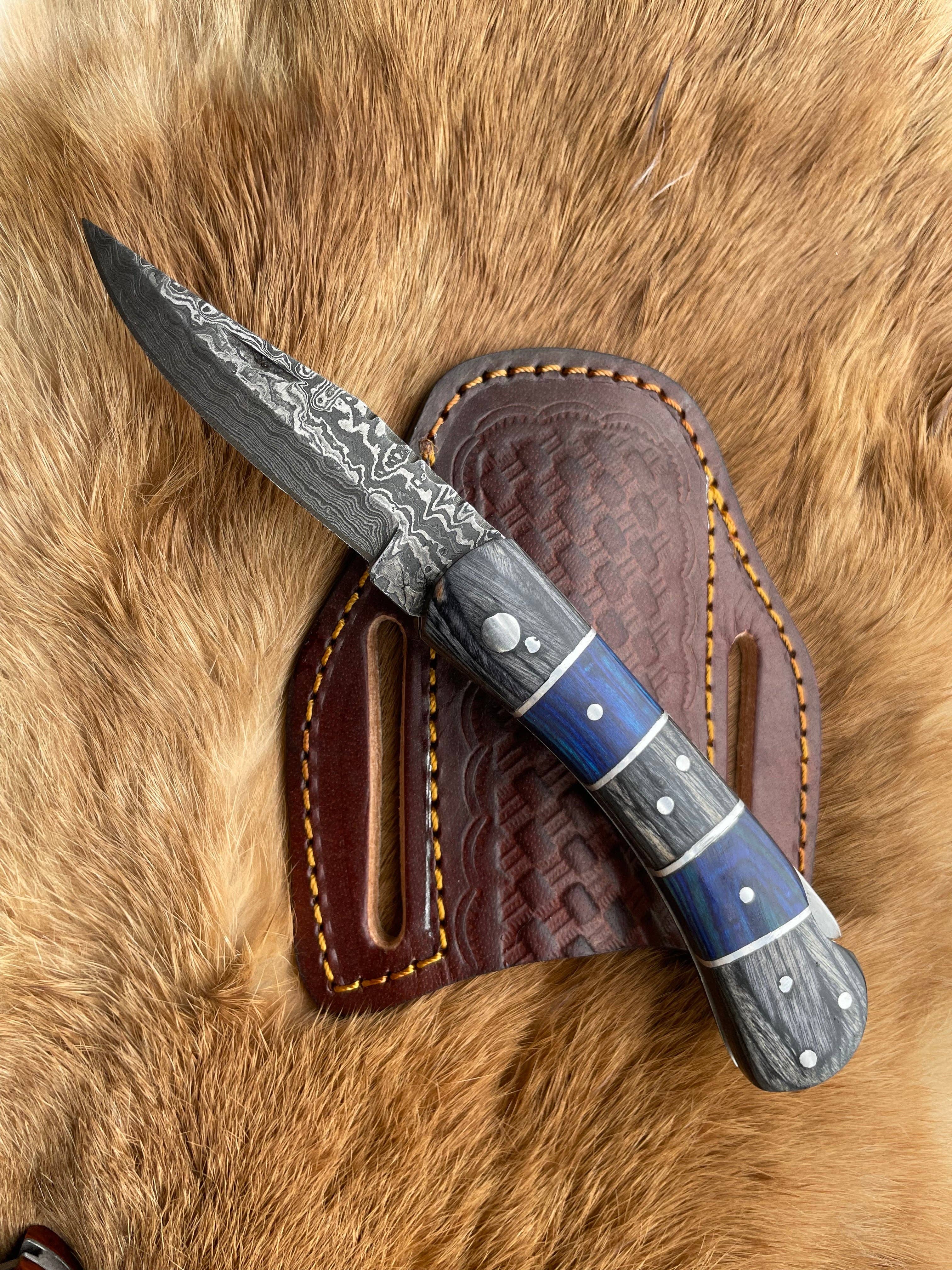 The Grey and Blue Wood 6.5 Damascus Steel Folding Knife, featuring a patterned blade and a blue and grey handle, is shown resting on its leather sheath atop a fur surface.