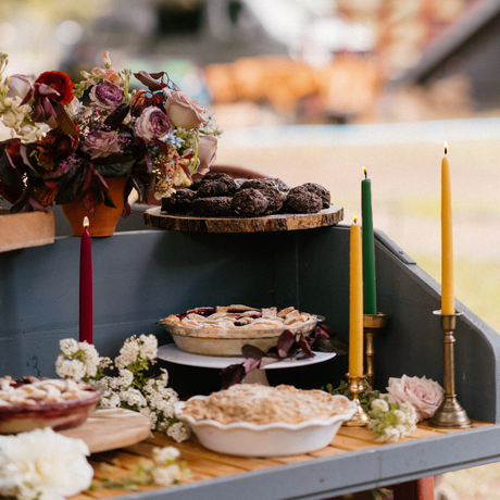 A variety of pies and pastries are showcased on a tiered stand alongside elegant floral arrangements, complemented by two eco-friendly, dripless, handmade 10 beeswax taper candles.