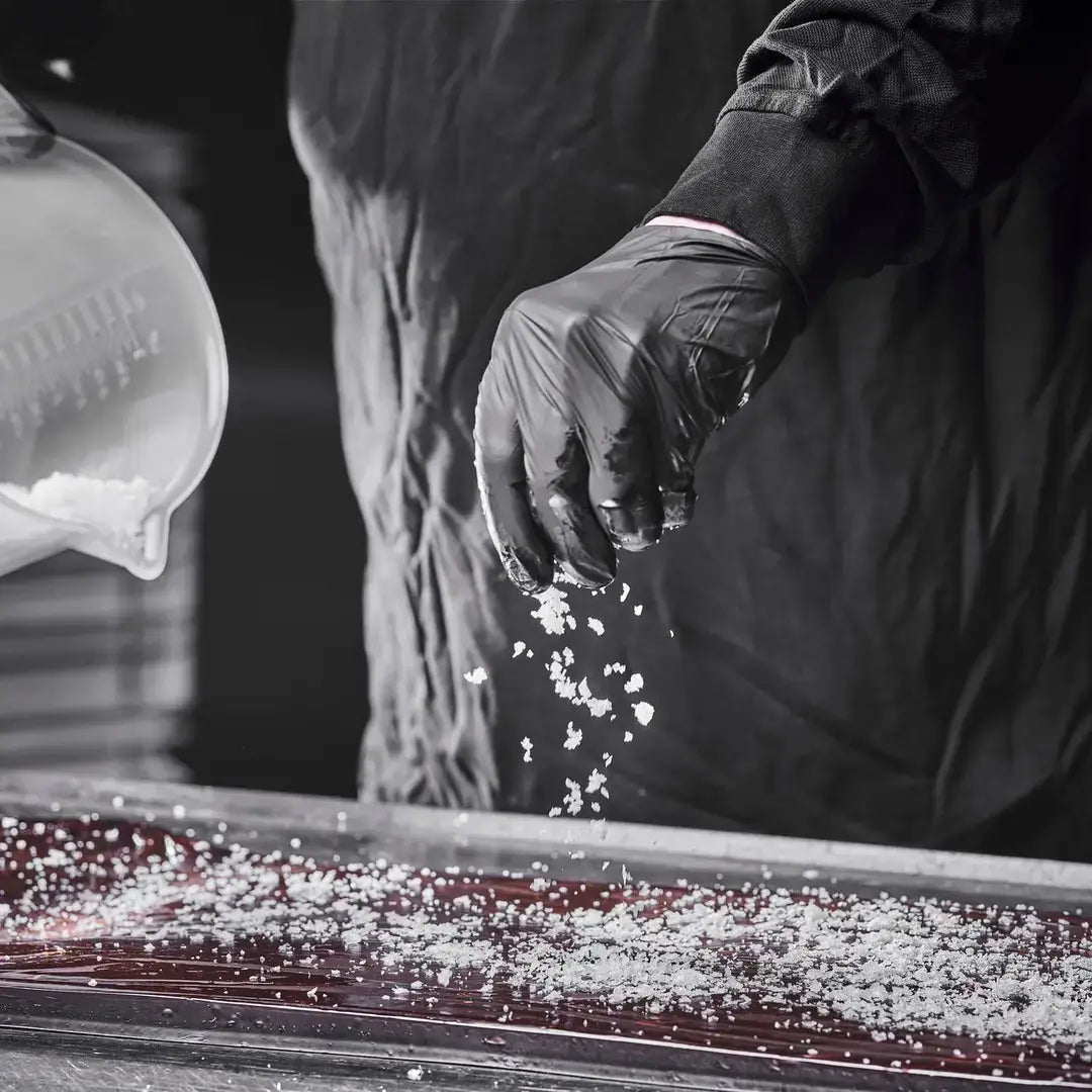 A person wearing black gloves sprinkles coarse salt over a flat surface, with additional salt visible in a container nearby.