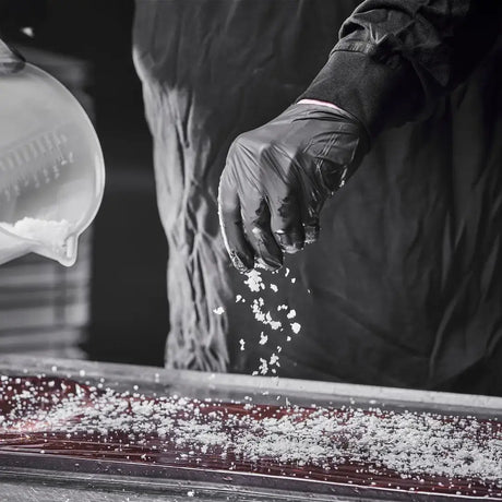 A person wearing black gloves sprinkles coarse salt over a flat surface, with additional salt visible in a container nearby.