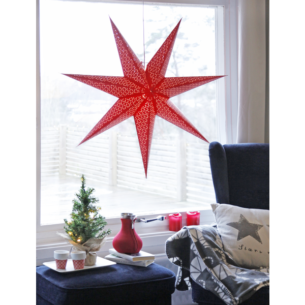 A decorative Light: Star Trading - Paper Dot Star in red, measuring 21.3" and made in Sweden, hangs by the window. Nearby, a small decorated tree enhances the cozy atmosphere, accompanied by a red teapot, cups, and a blanket on the sofa. Three candles flicker warmly on the windowsill.